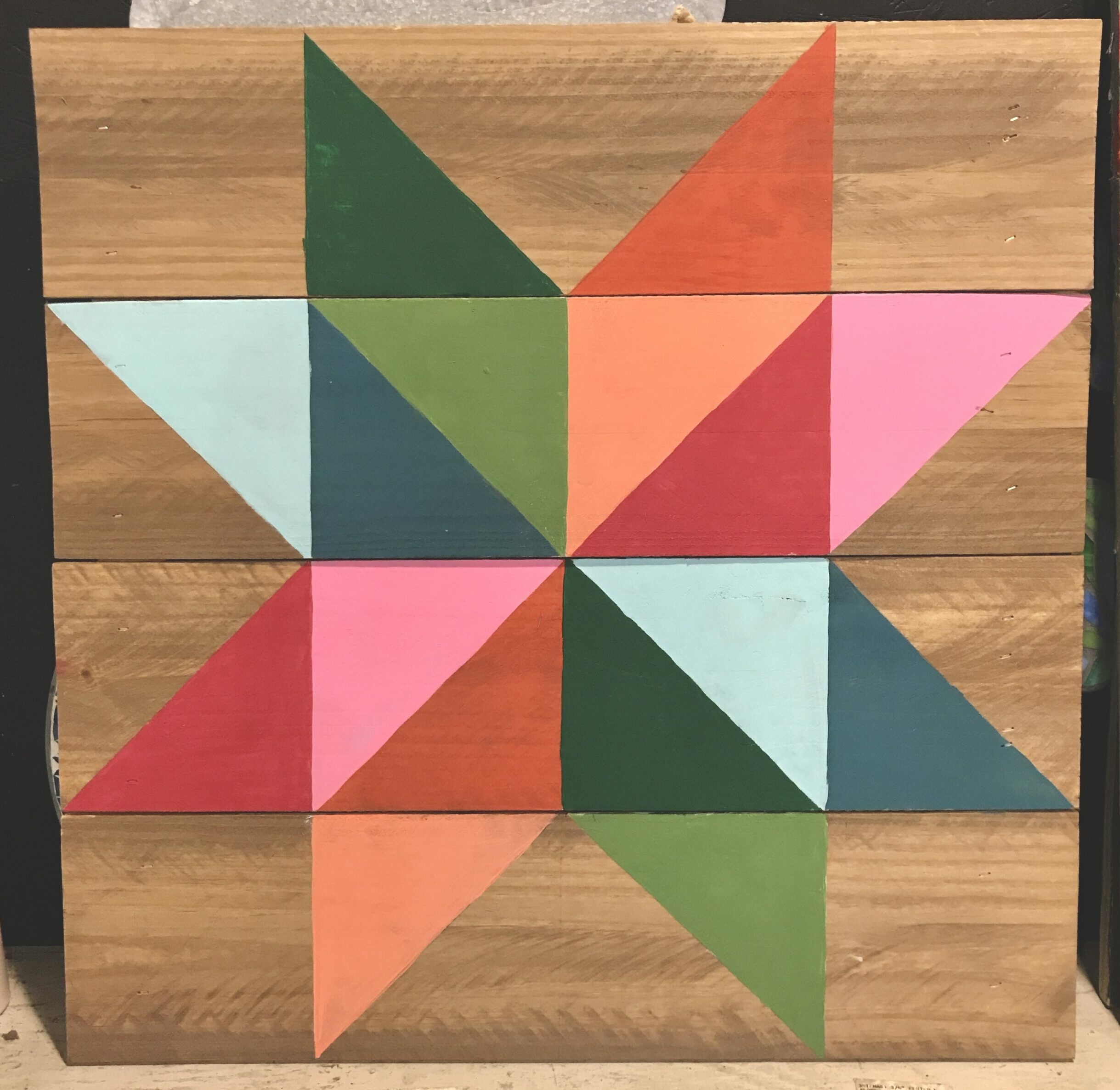 A wooden sign featuring green, red, pink, and orange paint that reveals a quilt-like pattern.